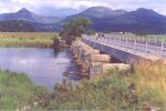 bridge piers across wide river; road bridge beside disused railway; mountain panorama beyond