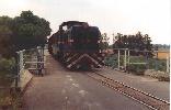 dirty blue locomotive with short train running along rails set into the road across a river bridge